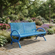 A bright blue slatted outdoor park bench with curved armrests and decorative metal legs, placed on a paved walkway in a landscaped garden with flowers, trees, and a lamp post.