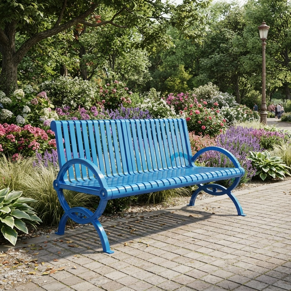 A bright blue slatted outdoor park bench with curved armrests and decorative metal legs, placed on a paved walkway in a landscaped garden with flowers, trees, and a lamp post.