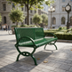 A green metal park bench with curved armrests and slatted back and seat, placed on a paved plaza in an urban setting with buildings, trees, and a clock in the background.