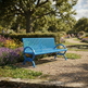 A bright blue slatted park bench sits on a gravel path in a landscaped garden with trees, flowers, and distant people walking along a path.
