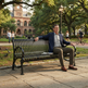A man in a navy blazer and khaki pants sits on a black metal park bench in a campus setting, with a historic brick building, trees, and other people in the background.