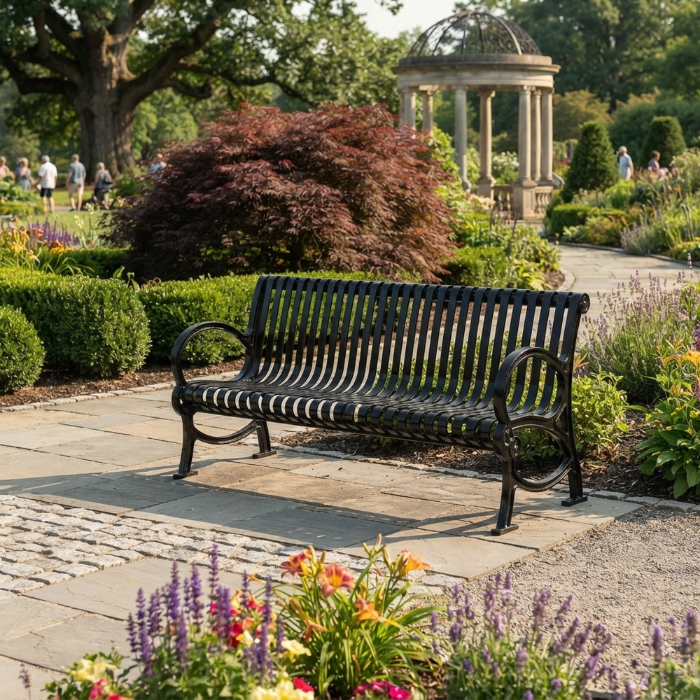 A black metal garden bench with curved armrests sits on a stone pathway in a landscaped park with flowers, shrubs, trees, and a pavilion in the background.