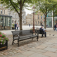A cobblestone plaza with two black metal park benches, leafy trees, a vintage-style lamppost, and historic stone buildings in the background. A man sits on one bench reading a newspaper, while others walk nearby.
