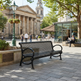 A black metal slatted park bench in a paved public square, with a stone classical building, trees, a lamp post, and a cafe in the background. A person sits on a bench at the cafe while another person walks nearby.