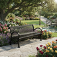 A black metal garden bench with curved armrests sits on a stone pathway in a lush, colorful garden with flowers, trees, and a pond.