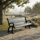 A modern park bench with dark metal arms and legs and light gray slatted seating, situated on a stone pathway in a landscaped park with a pond, trees, and distant city skyline.