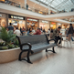 A black slatted public bench sits in the foreground of a busy multi-level shopping mall food court, with people seated at tables and storefronts including Taco Fusion and Asian Noodles in the background.