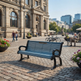 A blue slatted outdoor bench with black curved arms and legs sits on a cobblestone plaza in front of a large historic stone building. Pedestrians, bicycles, flowers, and a seated person are visible in the background.