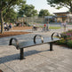 A modern dark metal park bench with curved backrests is shown in a landscaped public park with paved walkways, trees, a playground, a basketball court, and nearby buildings.