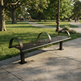 A dark metal park bench with curved backrests sits on a concrete sidewalk in a sunny park with green grass, trees, and a few distant people walking.