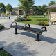 A modern black metal park bench with two curved backrests, situated on a paved outdoor plaza in a landscaped community center area with grass, trees, flowers, and nearby structures.