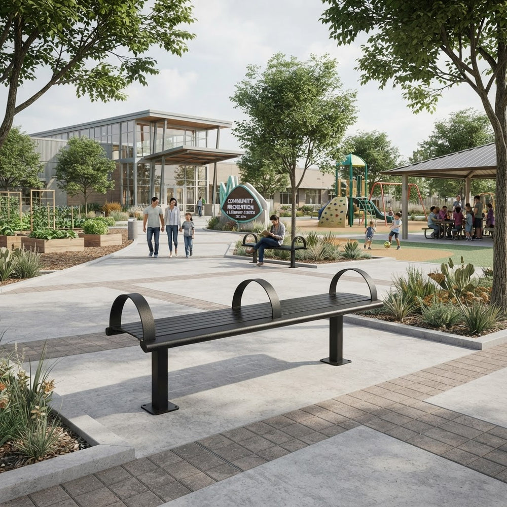 A modern black metal park bench with curved armrests situated on a paved walkway in a landscaped public park. Trees, planters, a playground, and a building are visible in the background, with a few people walking and sitting nearby.
