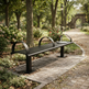 A modern black metal park bench with curved armrests sits on a stone-paved path in a lush, tree-lined park. A newspaper rests on the back of the bench, and the background features a stone archway, lampposts, and blooming flowers.