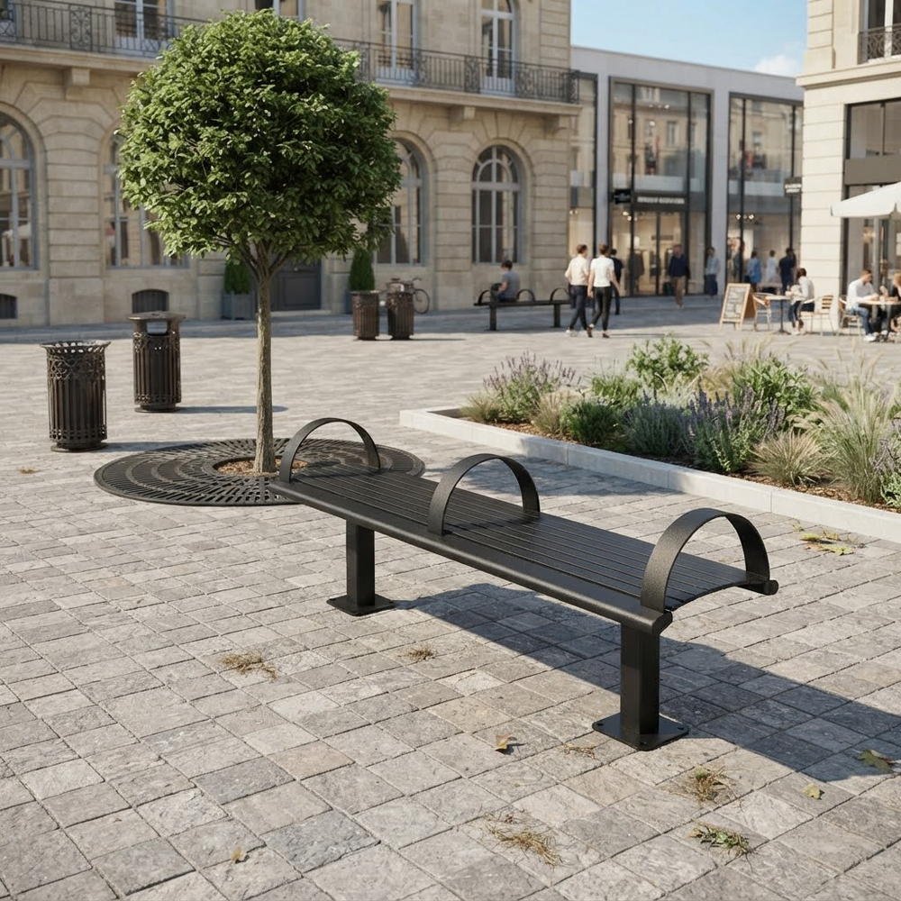 A modern black metal park bench with curved armrests sits on a stone plaza in front of a European-style building with arched windows and a small tree planted in a circular planter.