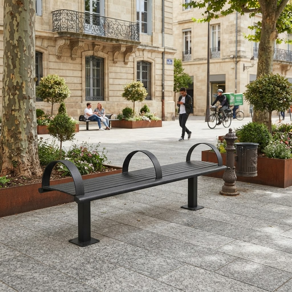 A modern black metal park bench with curved backrests is shown in an outdoor urban plaza with stone buildings, trees, pedestrians, and cyclists in the background.