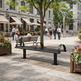 A modern black metal and wood park bench is shown in a real outdoor plaza with people walking, trees, flower boxes, and buildings in the background.