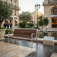 A modern wooden park bench with silver metal arms and legs, situated in a luxurious outdoor plaza next to a fountain and a marble information plaque.