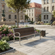 A modern outdoor park bench with dark brown wooden slats and silver metal arms and legs, situated in a paved public square with flowers, trees, and people in the background.