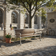 A wooden park bench with a silver metal frame sits on a cobblestone plaza in front of a stone building, with a tree, lamp post, small table, and bicycle nearby.