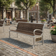 A modern outdoor park bench with brown wooden slats and a silver metal frame, situated on a cobblestone plaza in a city setting with people sitting on other benches in the background.