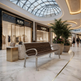 A modern metal and wood bench sits in a luxury shopping mall atrium with marble floors, large potted plants, and storefronts including AURA and LOMÈRE.