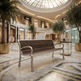 A modern outdoor-style bench with a dark brown slatted seat and silver metal frame, placed in a luxurious, upscale shopping mall atrium with marble floors, palm trees, and Gucci storefronts visible in the background.