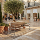 A wooden park bench with curved silver metal arms and legs is placed in a sunlit outdoor shopping plaza with a large potted tree, stone paving, storefronts, and a few pedestrians in the background.