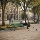 A man sits on a green metal park bench in a paved town square, reading a book. Trees, historic stone buildings, other pedestrians, and pigeons are visible in the background.