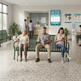 A bright, modern hospital or clinic waiting area with several people seated on green benches. In the foreground, an elderly woman uses a walking cane, a man reads a magazine, and a young woman checks her phone.