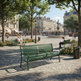 A green metal park bench in the foreground of a sunny urban plaza with cobblestone ground, trees, benches, trash cans, and historic buildings in the background. A seated person is visible on a bench in the plaza.