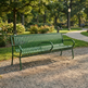 A green metal park bench with curved armrests sits on a gravel path in a sunny, tree-lined park with a winding pathway and flowers in the background.