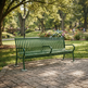 A green metal park bench with curved armrests sits on a stone-paved area in a sunny park with grass, trees, and flower beds in the background.