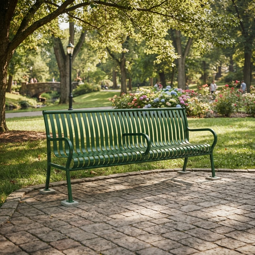 A green metal park bench with curved armrests sits on a stone-paved area in a sunny park with grass, trees, and flower beds in the background.