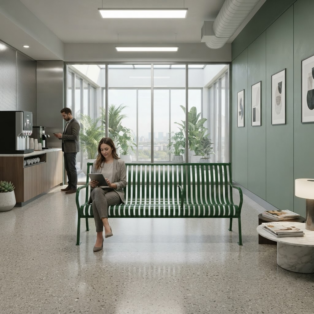 A woman sits on a green metal bench in a modern office lobby, using her laptop. A man stands at a coffee station in the background near large windows and indoor plants.