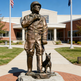 Bronze statue of a person in uniform with a dog on a pedestal in front of a building.