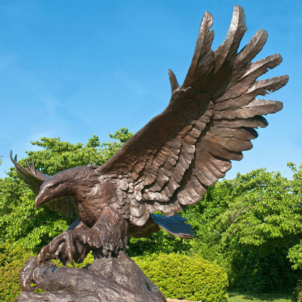Bronze statue of an eagle in flight with a clear blue sky and green trees in the background
Bronze raptor sculpture perched on a rock with powerful open‑wing stance