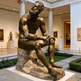 Bronze statue of a seated figure in a museum setting with columns and other sculptures.