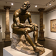 Bronze statue of a seated figure in a museum setting with classical columns and artwork.