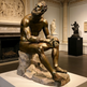 Bronze statue of a seated figure in a museum setting with other sculptures in the background.