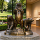 Bronze statue of three bears on a pedestal with a garden and building interior in the background.