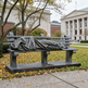 Statue of a person draped in a blanket on a bench in an urban park setting.
Realistic bronze sculpture of a figure lying under a blanket on a bench