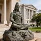Bronze statue of a seated figure on a rock with a building in the 
background
Sculpture of Shiva in a cross‑legged pose with detailed jewelry and topknot