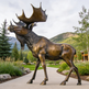 Bronze moose statue in a natural setting with trees and mountains.