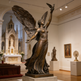 Bronze statue of an angel holding a dove in a museum setting with religious architecture in the background.