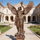 Bronze statue of an angel holding a torch in front of a classical building.
Sculpture of a winged woman lifting a torch on pedestal