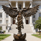 Bronze statue of an angel with wings spread in front of a classical building

Sculpture of a winged female figure emerging from a broken sphere with radiant crown