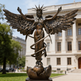 Bronze statue of an angel with wings and a snake around her waist in front of a building.
Detailed winged female statue with crown and upward‑reaching pose
