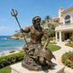 Bronze statue of a bearded man with a trident on a pedestal in front of a luxurious house by the ocean.