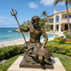 Bronze statue of a bearded figure with a trident on a turtle, set against a luxurious beachfront property.