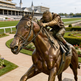 Bronze statue of a jockey on a horse in front of a racecourse building.